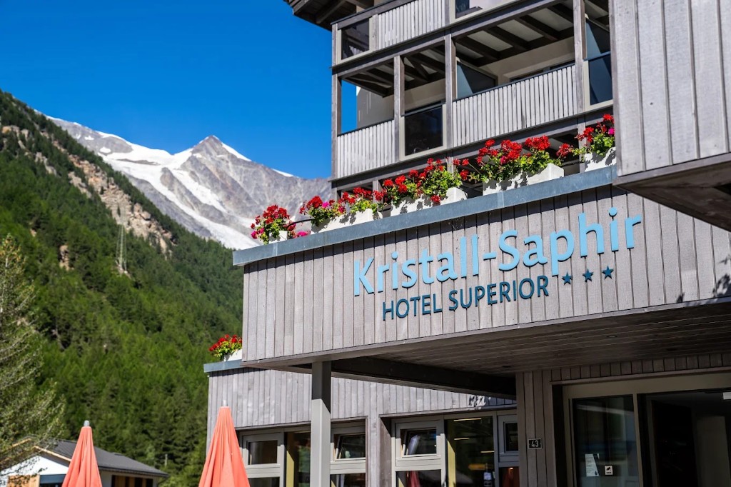 Exterior of Hotel Kristall-Saphir Superior in Saas-Almagell with wooden balconies, red geraniums and alpine peaks behind