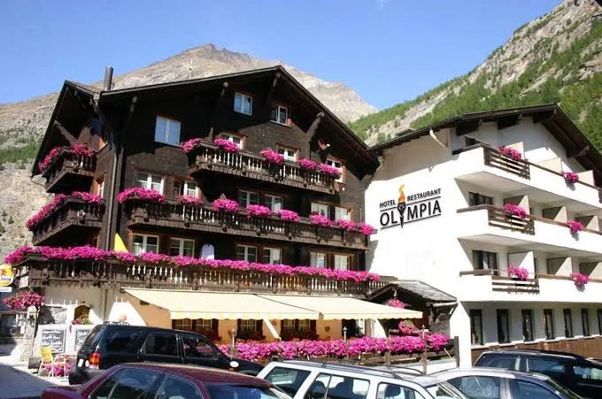 Hotel Restaurant Olympia in Saas-Almagell with wooden and white facades, pink flower balconies and a mountain backdrop