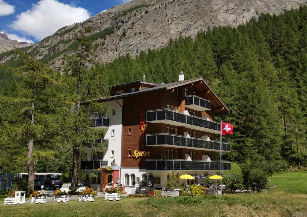 Exterior of Hotel Sport in Saas-Almagell with wooden balconies, Swiss flag and forested slopes behind